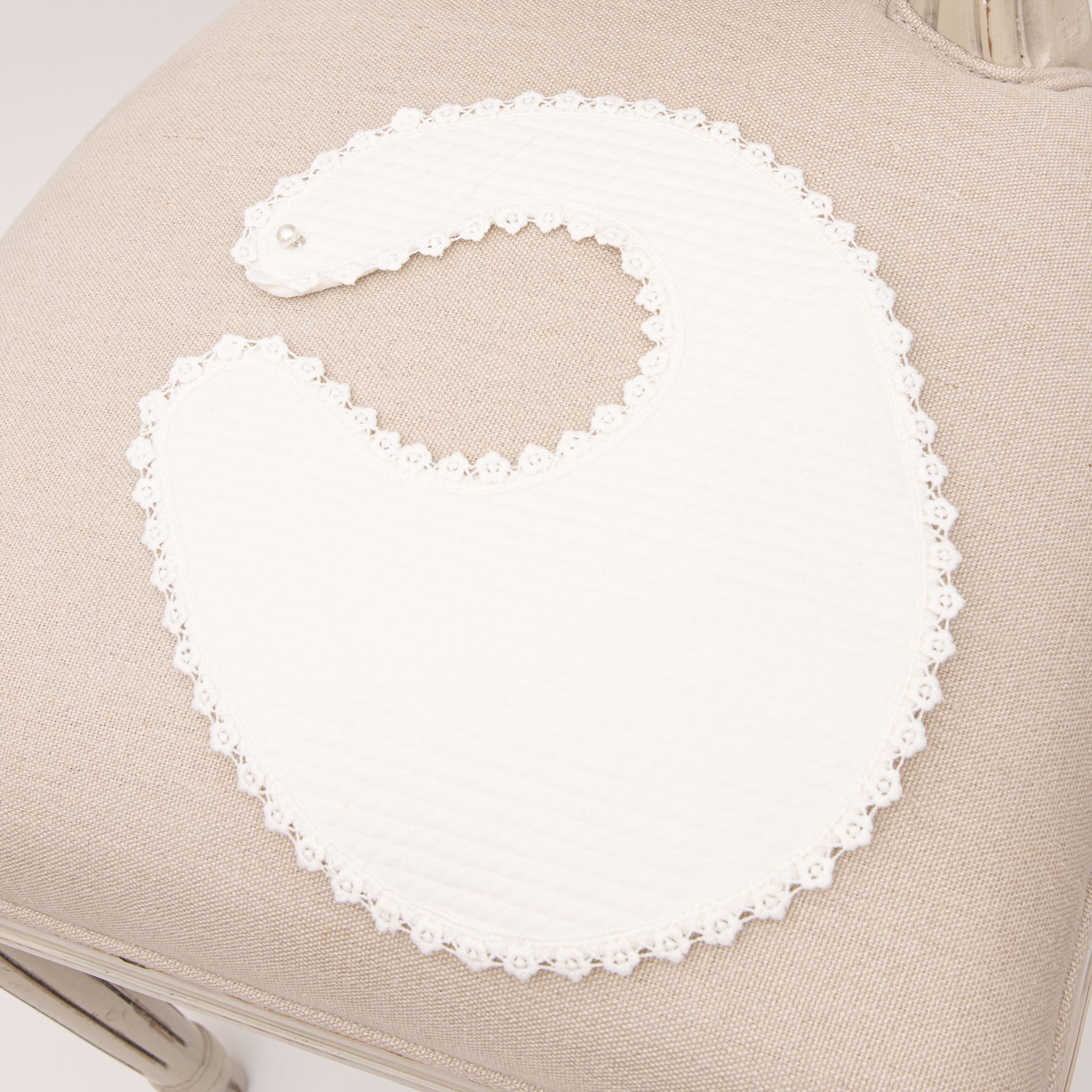 The Rose Accessory Bundle - Save 15% includes a white baby bib with a lace trim and matching lace baby shoes, elegantly displayed on a beige cushioned chair. The bib's scalloped edge and intricate detailing make it an exquisite addition to any baptism accessory set.
