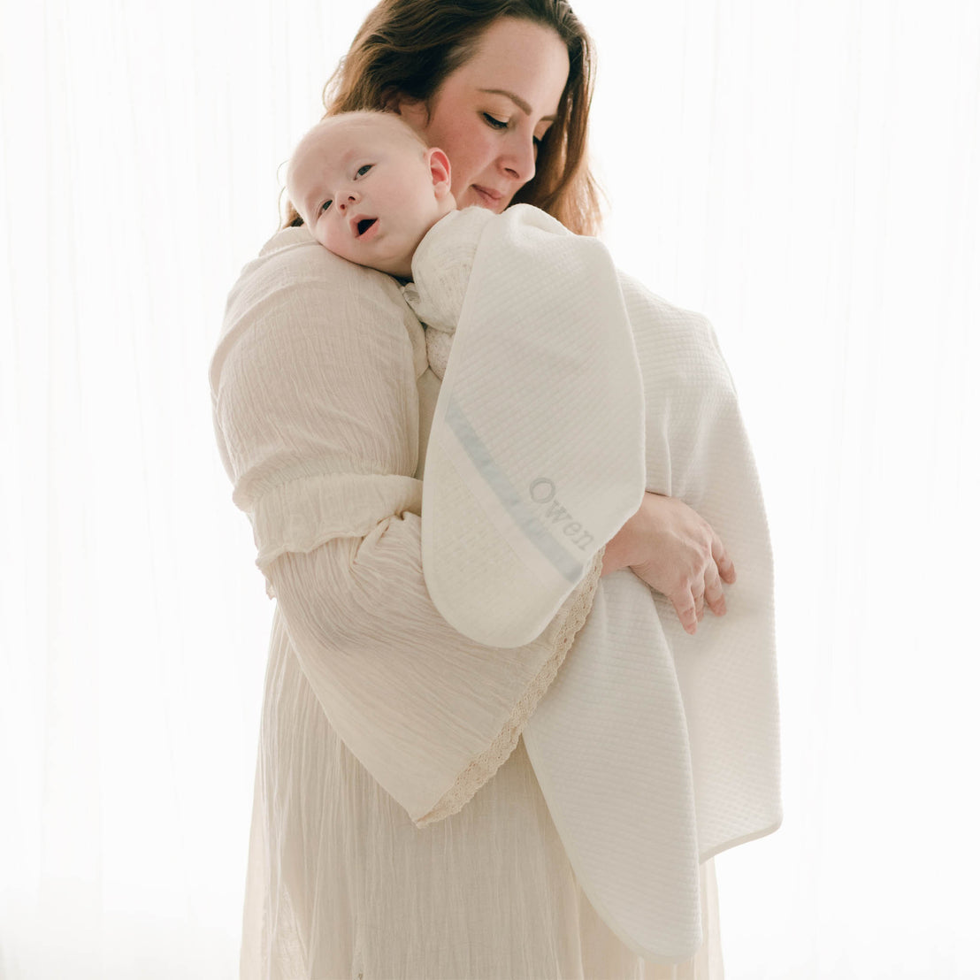 Photo of a mother and her baby wrapped in the Owen Personalized Blanket. The corner features an ivory knit and the name "Owen" embroidered with light blue thread
