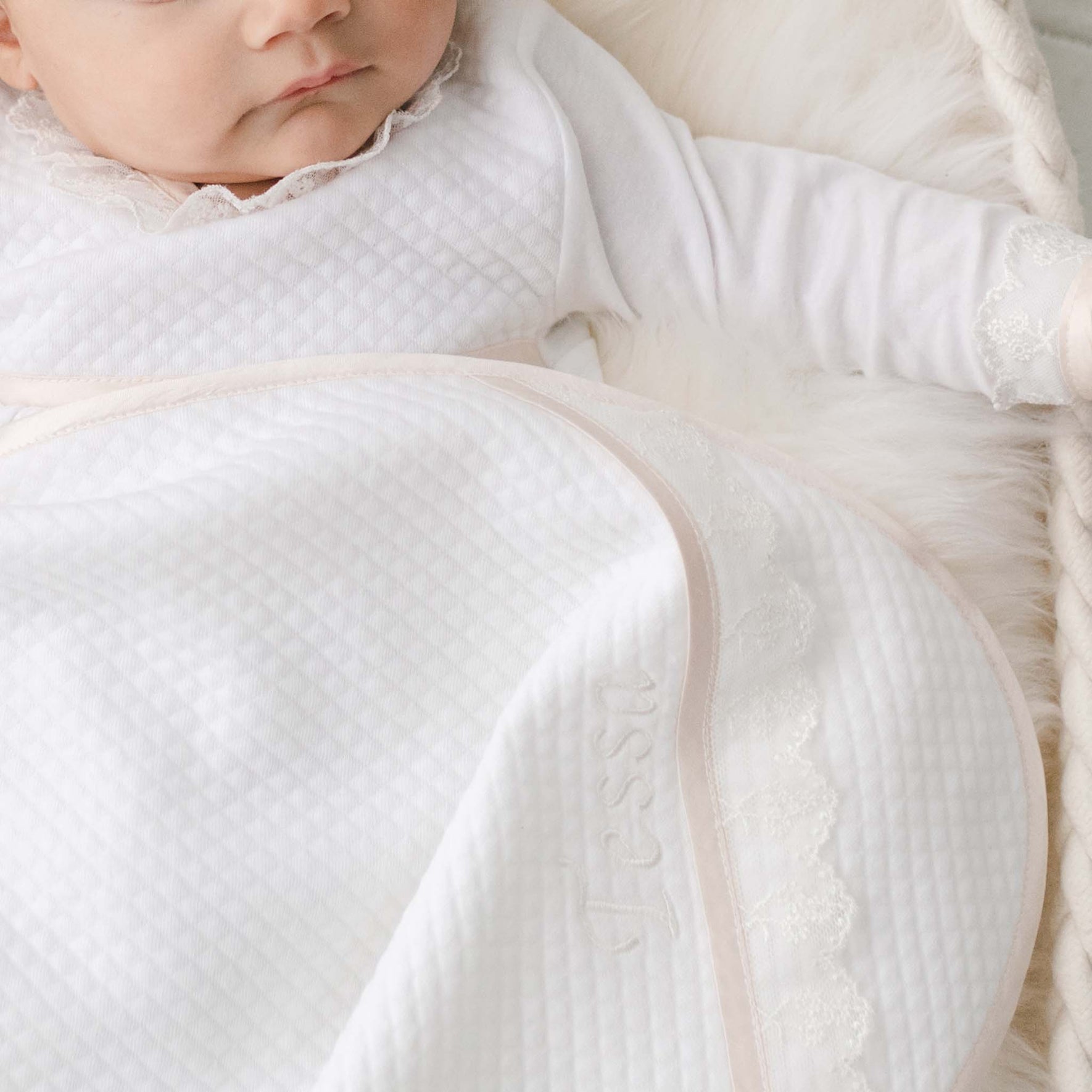 Newborn baby girl with the Tessa Personalized Blanket made from a textured cotton in white and featuring a ivory embroidered lace trim and personalized name embroidery. 