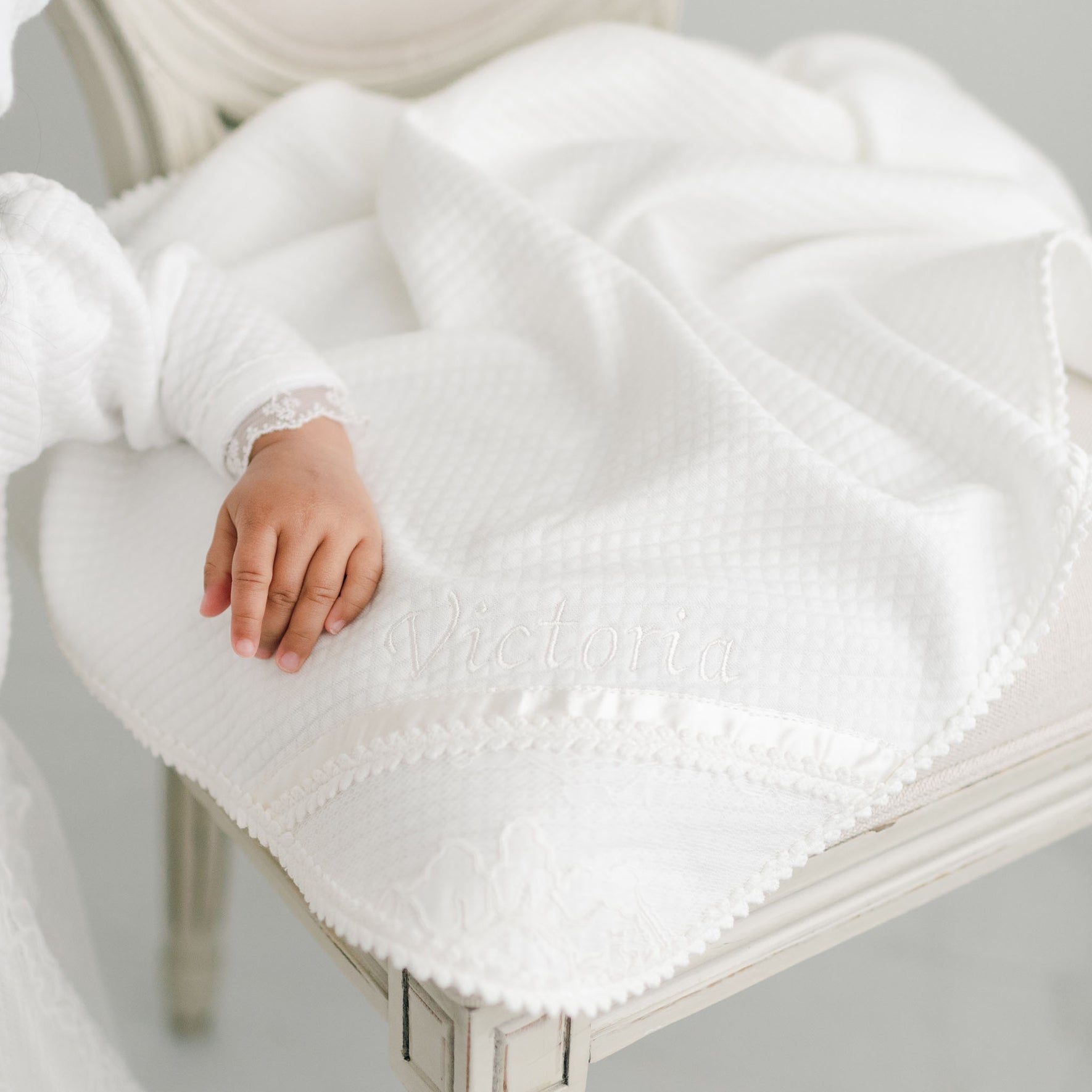A baby's hand gently lies on a soft, white Victoria Personalized Blanket draped over a chair, crafting an ideal setting for a special gift. The exquisite christening blanket is beautifully embroidered with the name "Victoria," while the baby's sleeve showcases delicate lace trim.