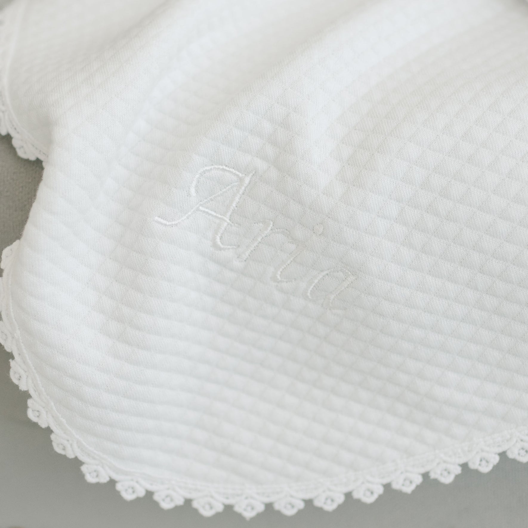 The Aria Personalized Blanket is a white textured fabric with "Aria" embroidered in matching white thread. Featuring a diamond pattern and delicate lace trim along the edge, it's perfect as a christening blanket and makes an ideal personalized gift.