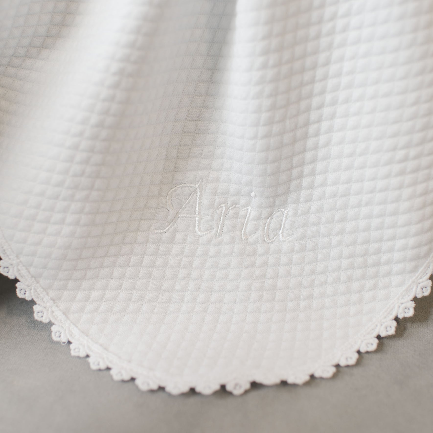 A detailed view of the Aria Personalized Blanket reveals its white, quilted texture and scalloped edges. The name "Aria" is elegantly embroidered in the center with white thread, making it an ideal personalized gift for a baptism outfit.