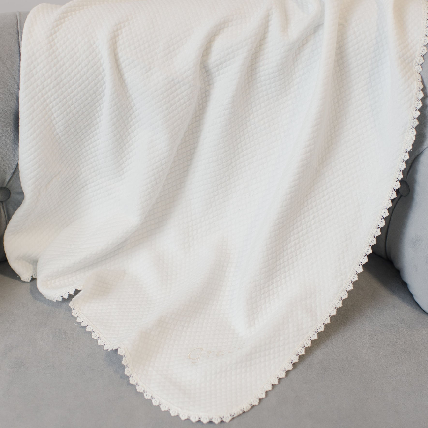 The Grace Accessory Bundle, featuring a personalized quilted blanket with a white waffle-pattern and lace trim, is elegantly draped over a gray sofa.