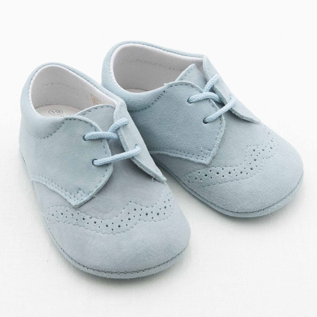 A pair of light blue Harrison Suede Shoes designed for baby boys, featuring handmade suede material with laces and decorative perforations reminiscent of brogue detailing. The shoes are positioned side by side on a plain white background.