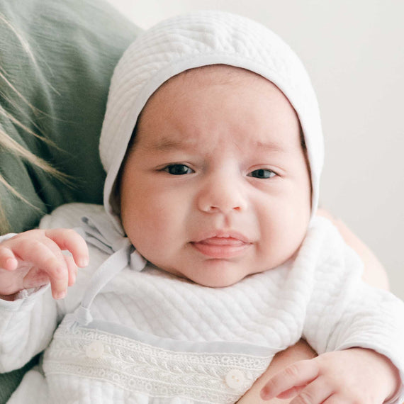 A close up detail of a newborn baby boy wearing a Harrison Quilted Bonnet made with soft quilted cotton in white and trimmed in a light blue linen
