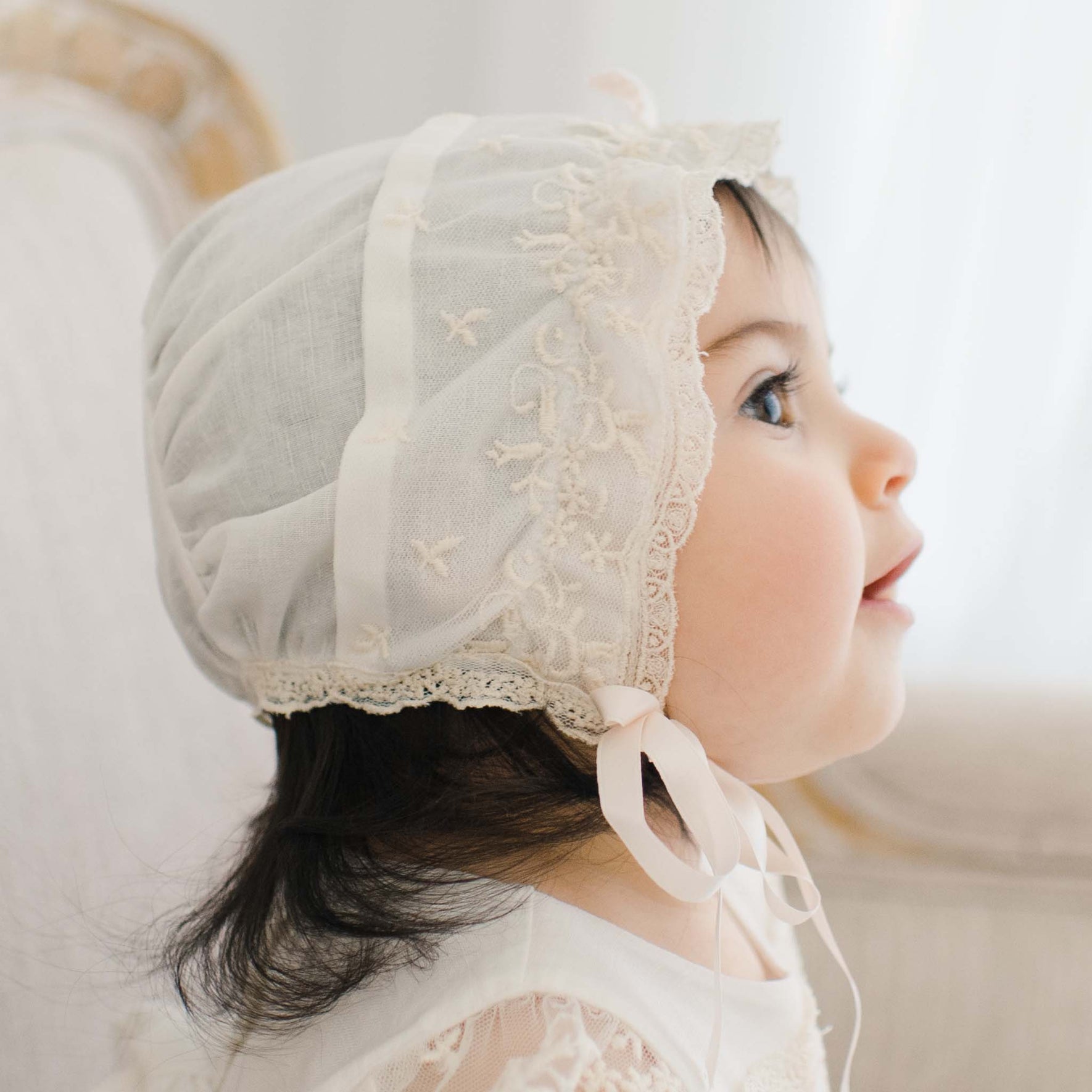 A young child is pictured in profile wearing a white Jessica Linen Bonnet with Champagne Lace details. The child's dark hair is partially visible under the bonnet, and an ivory ribbon ties under the chin. The background is softly blurred, focusing attention on the child's face and attire.