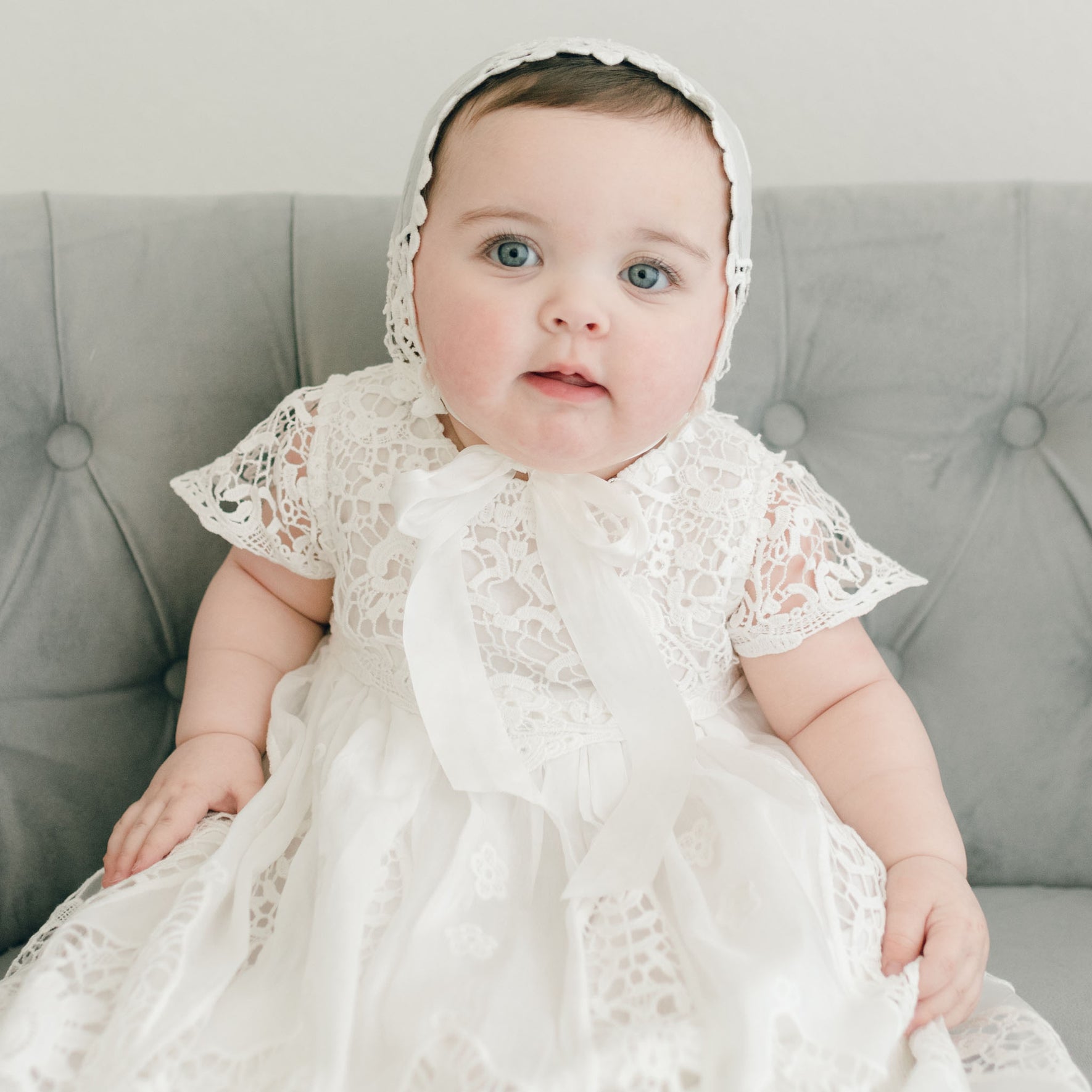 A baby wearing the Grace Christening Gown & Bonnet sits on a grey tufted couch. The intricate patterns on the outfit are complemented by a silk sash, and a white ribbon is tied at the neckline. The baby looks forward with a neutral expression.