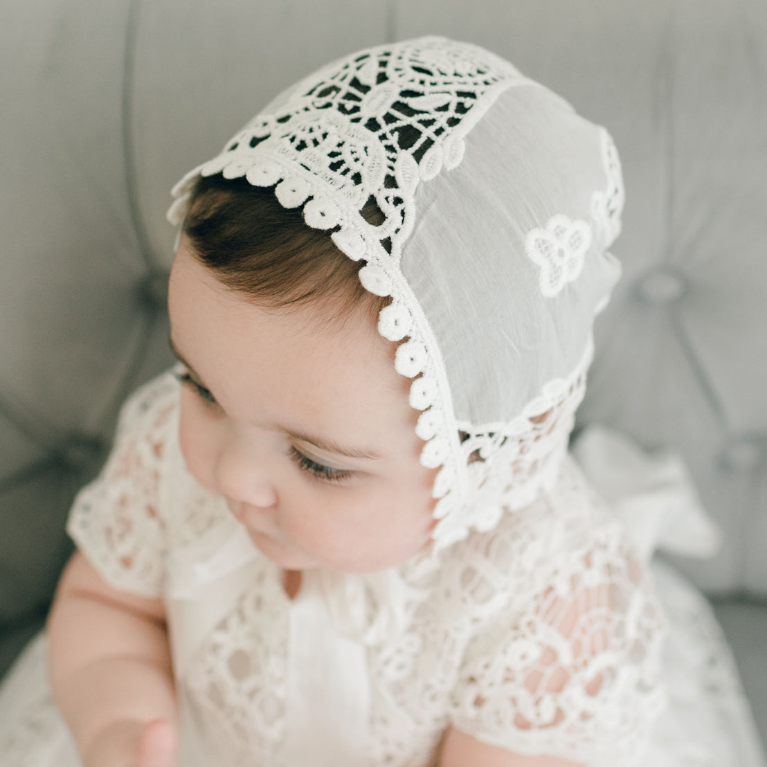 Sitting on a gray tufted chair, a baby adorns an ivory Grace Lace Bonnet paired with a matching lace outfit. The christening bonnet is detailed with intricate floral patterns and a scalloped edge. The baby's gaze is directed off to the side.