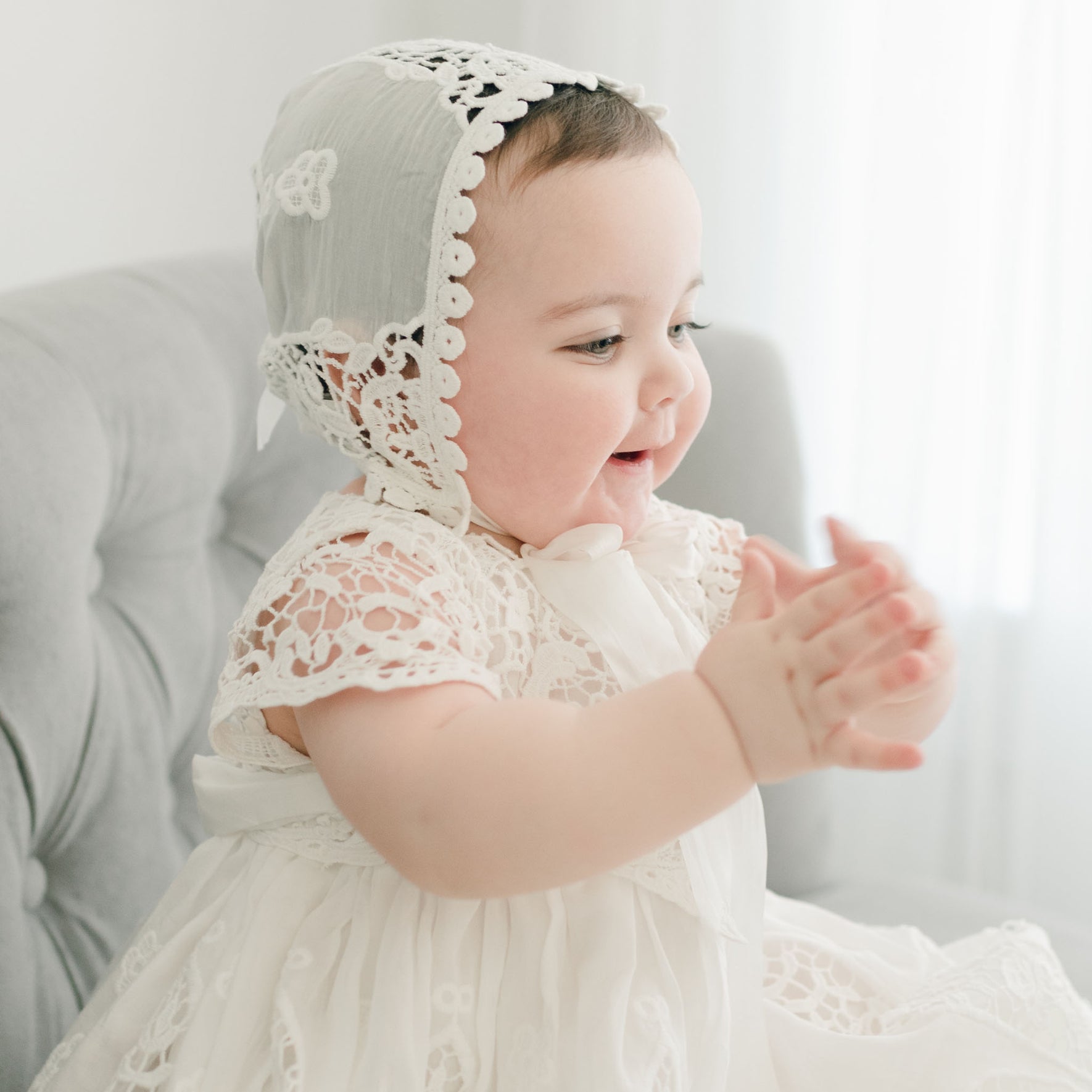A joyful baby wears the Grace Christening Gown & Bonnet while sitting on a light gray cushioned chair. The baby, with a gleeful expression, claps their hands and looks to the side. White curtains can be seen in the background.