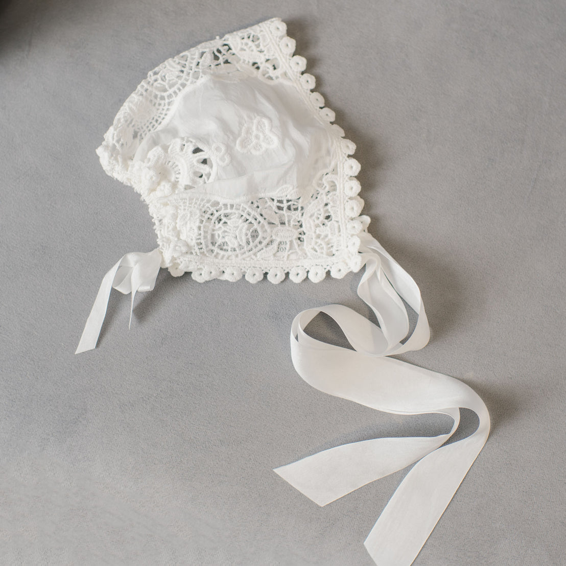 The Grace Lace Bonnet, an ivory baby bonnet featuring intricate lace embroidery and patterns, lies on a gray surface. This christening bonnet is adorned with decorative trim and has two long ribbon ties.