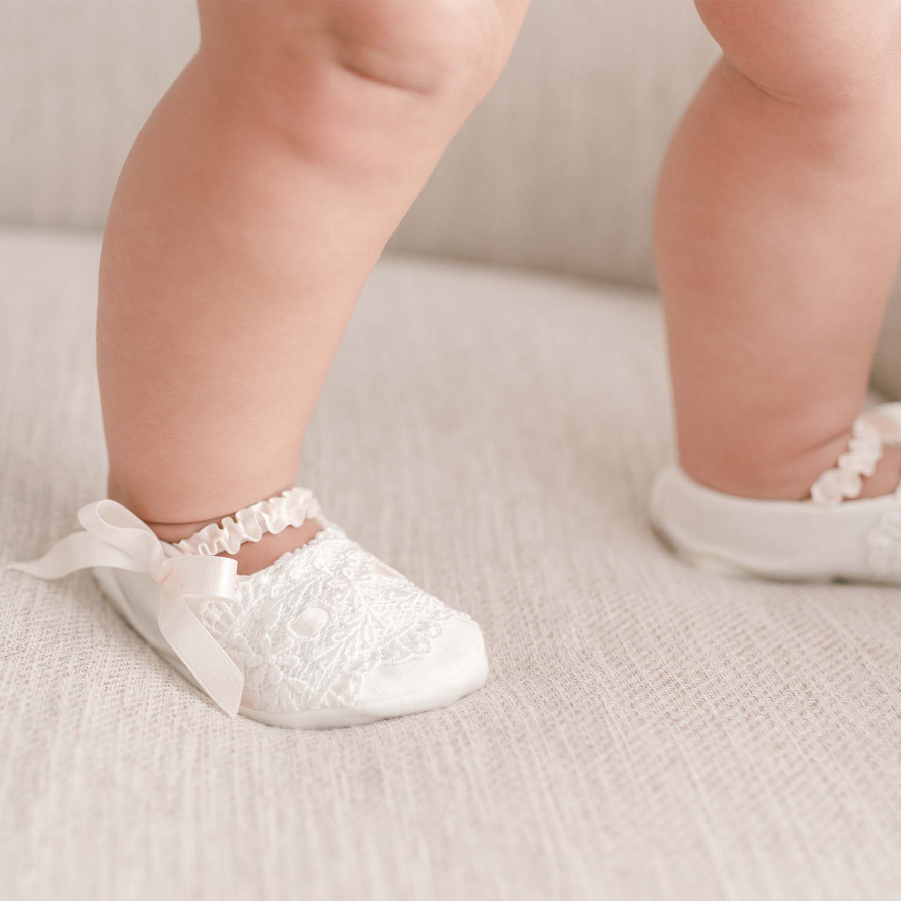 Close-up of a baby wearing the Emma Christening Booties, featuring ivory lace and adorned with pink silk ribbons, sitting on a light-colored fabric surface. The focus is on the baby's feet and booties.