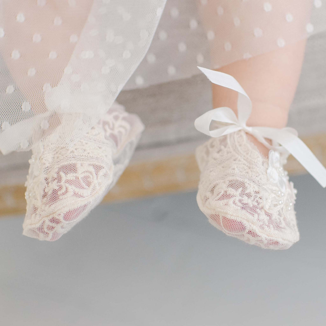 A close-up image of a baby's feet wearing delicate, Jessica Lace Booties adorned with pearl beading. The baby is also wearing a light, sheer, polka-dotted fabric, likely a dress or blanket, which can be partially seen in the background.