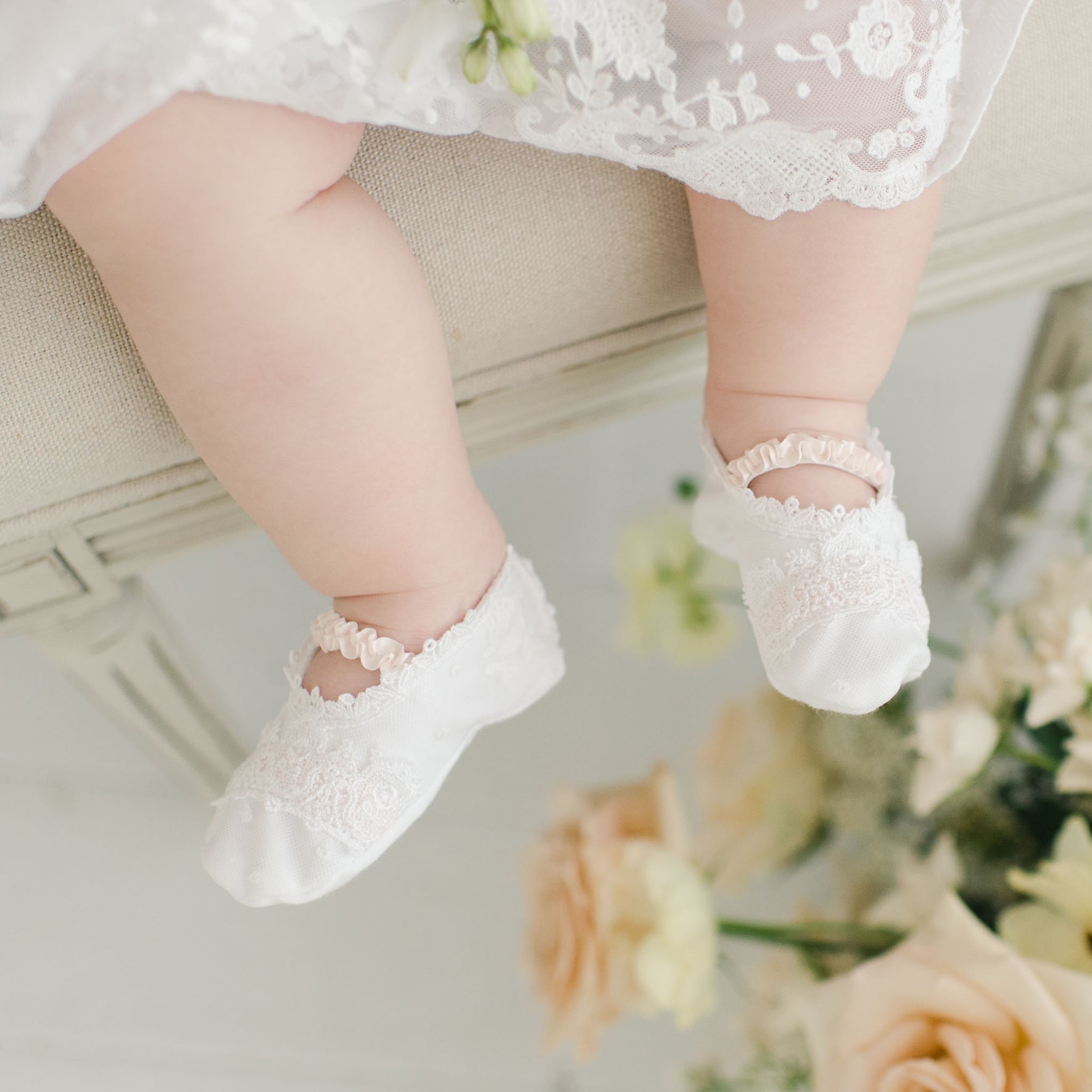 Close-up of a baby's legs wearing Melissa Christening Booties crafted from white Venice lace, seated on a light-colored surface. The baby is dressed in an elegant outfit made from 100% pima cotton. In the background, there are light-colored flowers, mainly white and soft peach roses.