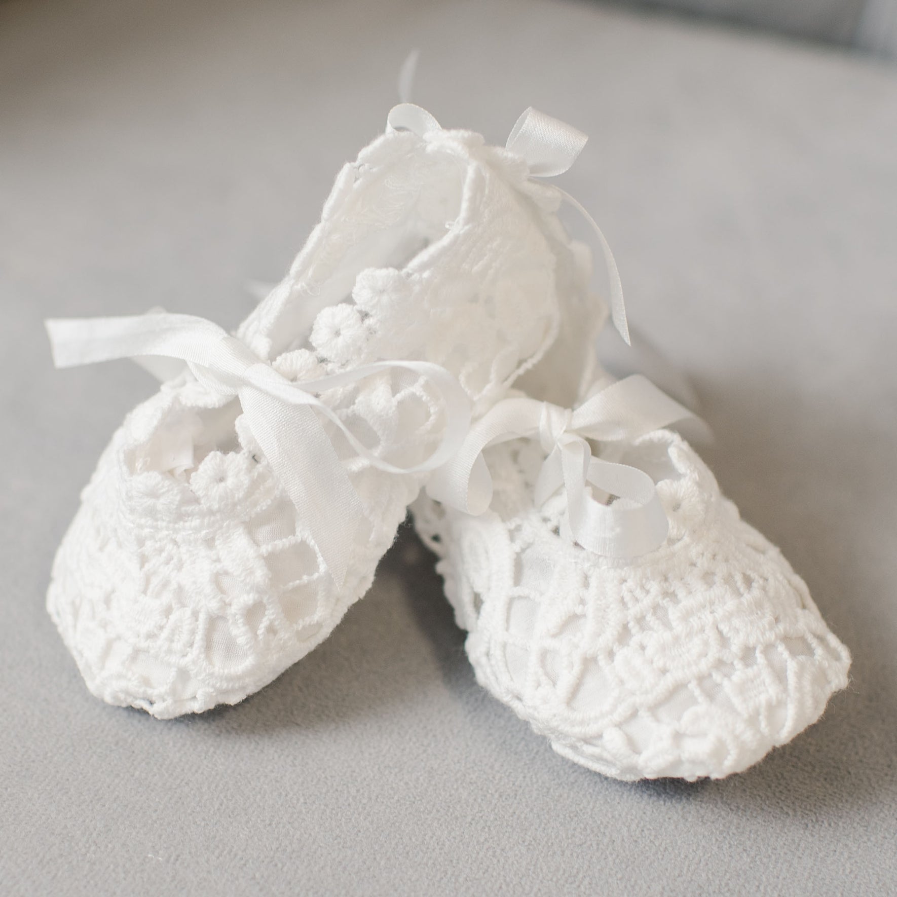 A pair of Grace Lace Christening Booties with ribbon ties are placed on a gray surface. These booties feature intricate patterns and a delicate design, perfect for completing a Christening Outfit.