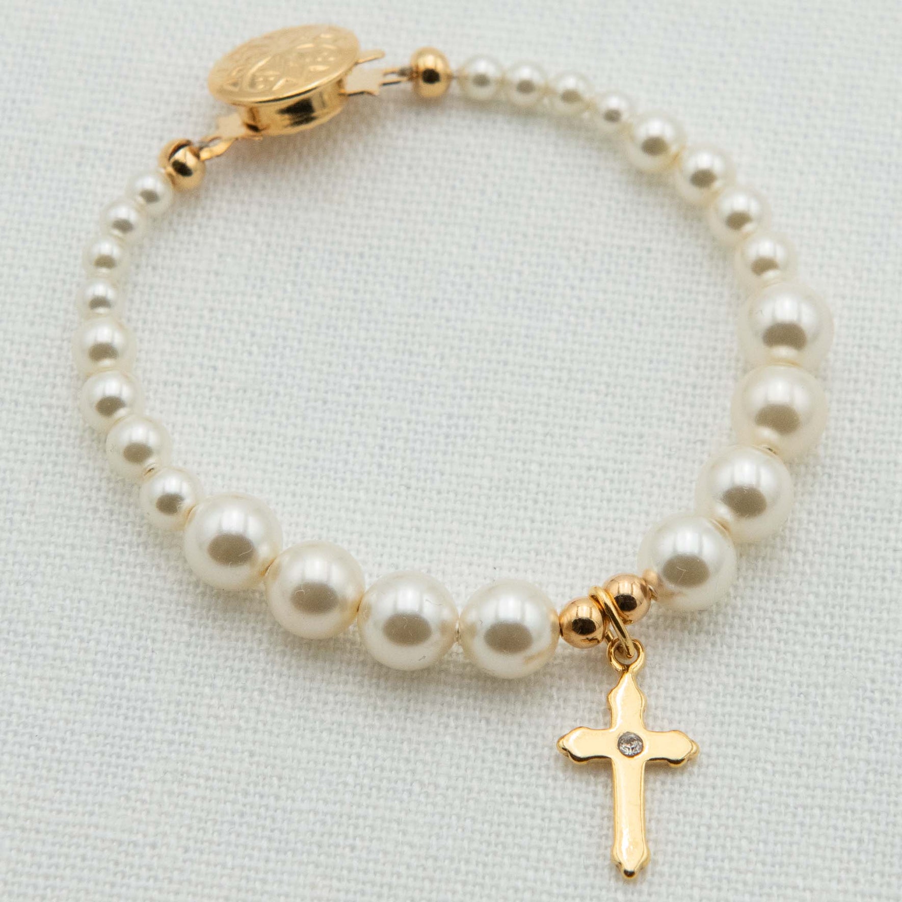 A pearl and gold bracelet featuring cream pearl beads, a gold clasp with a round medallion, and a dangling gold cross charm with a small clear stone, displayed on a textured white fabric background.