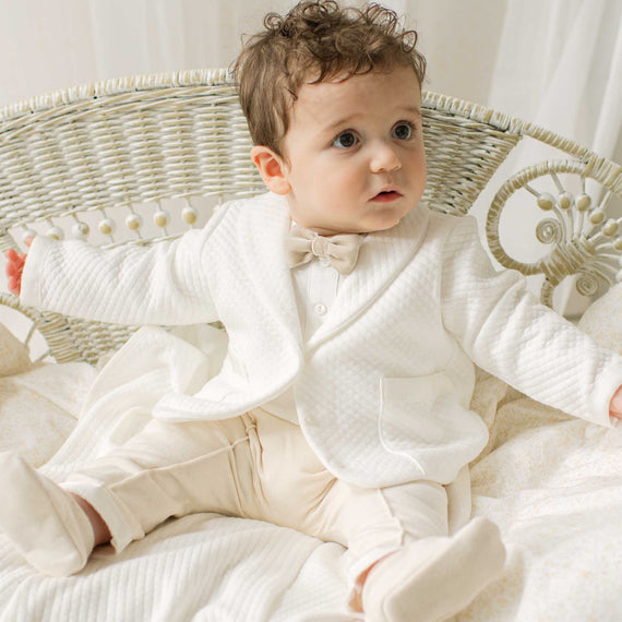 Baby boy in an ivory and tan outfit sitting in a wicker chair with a white background