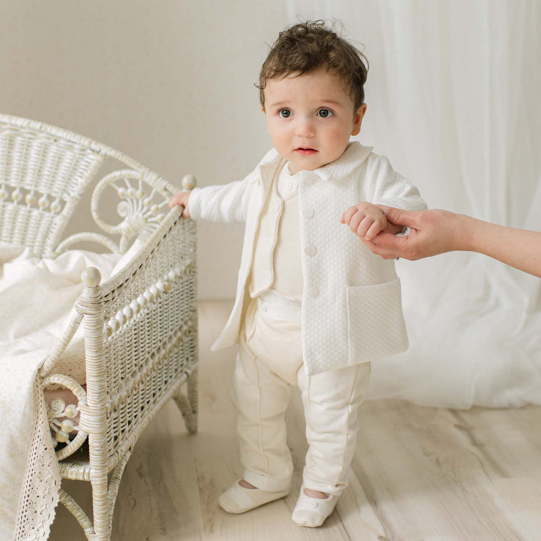 Baby boy in a Braden Coat Suit and matching Vest Suit standing next to a wicker chair in a bright room. Part of the Christening Gown Baby Boy Suit Collection.