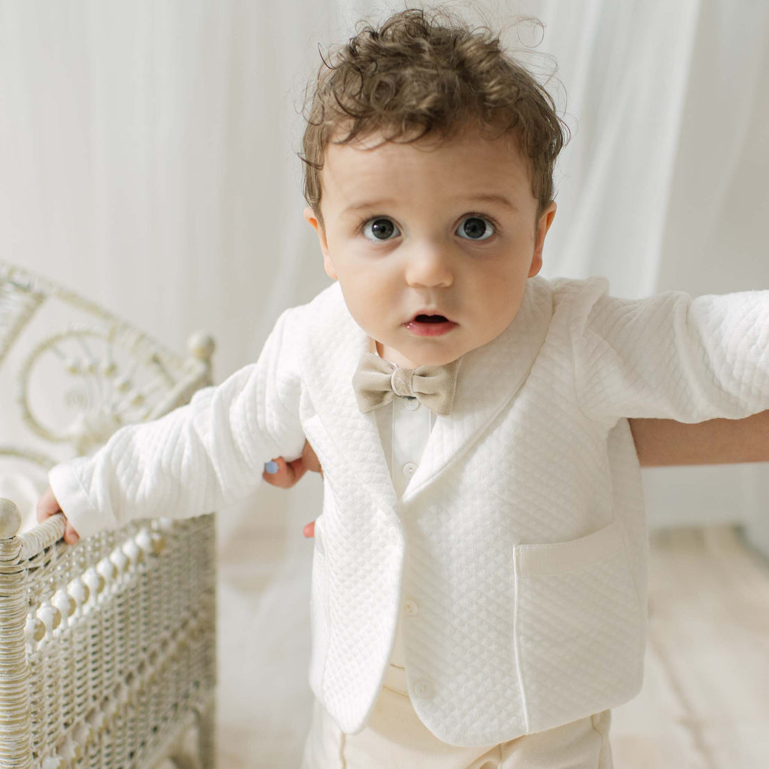 Baby in a ivory textured suit with a bow tie standing against a light background