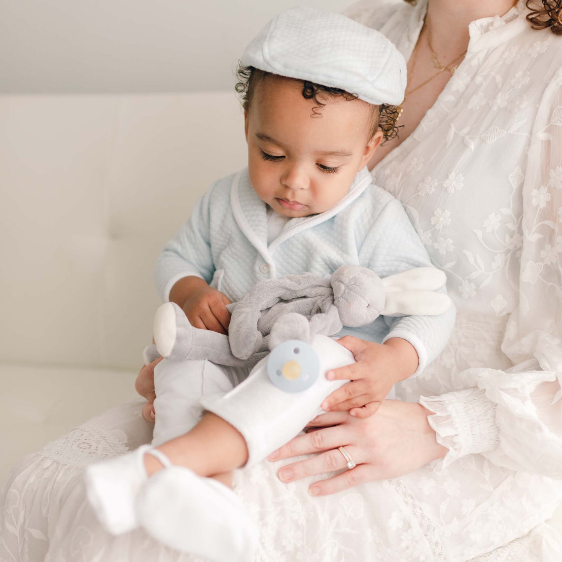 Baby boy sitting on a white couch with his mother and wearing the powder blue Asher 3-Piece Suit, including a folded collar jacket, white pants and a onesie with buttons (and matching Quilted Newsboy Cap). 
