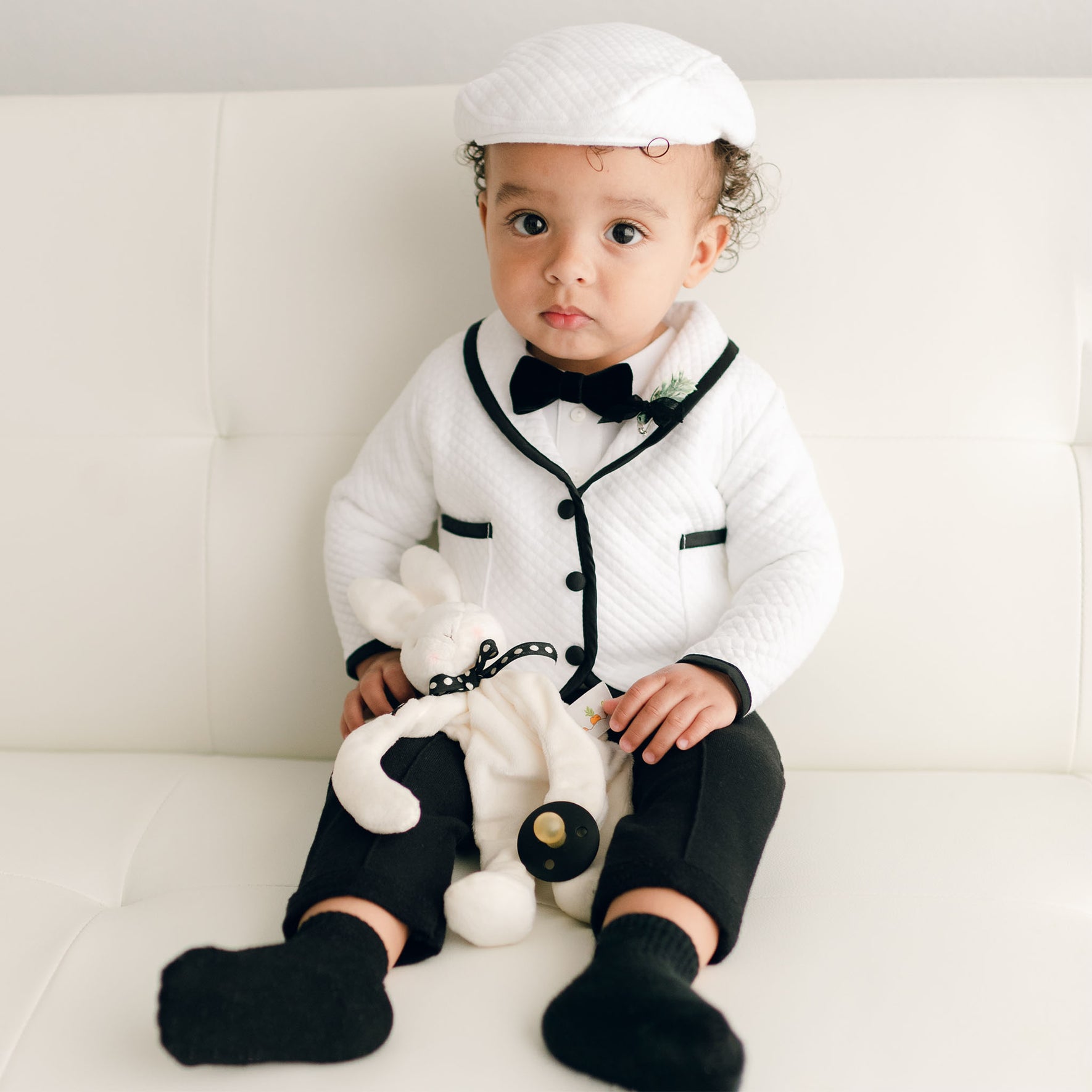 A baby sitting on a white couch in the James 3-Piece Suit, holding a plush bunny, with black pants and socks complementing the outfit.