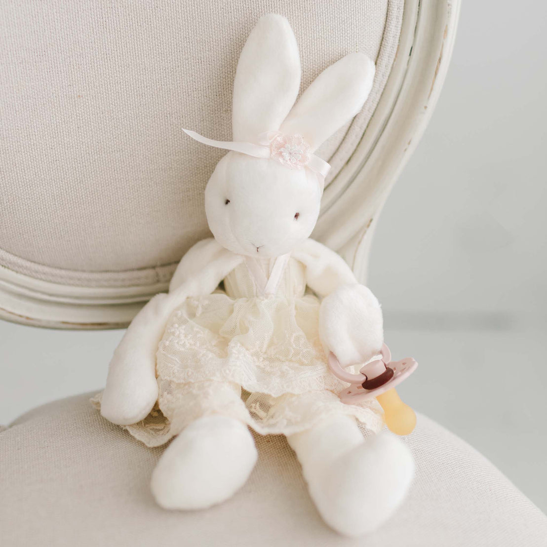 A white plush bunny toy wearing a lace dress with a pink bow on its head sits on an off-white upholstered chair. The bunny is clutching a Jessica Silly Bunny Buddy | Pacifier Holder.