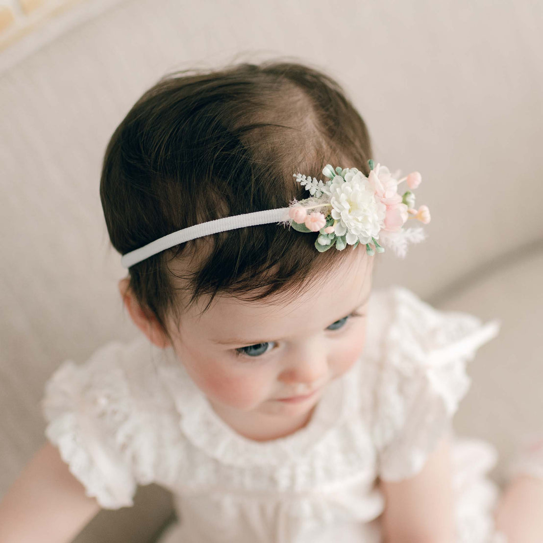 Top view of a baby girl in the Charlotte Flower Headband, adorned with pink and white flowers. She is standing on a linen chair, with her head turned and gaze lowered.