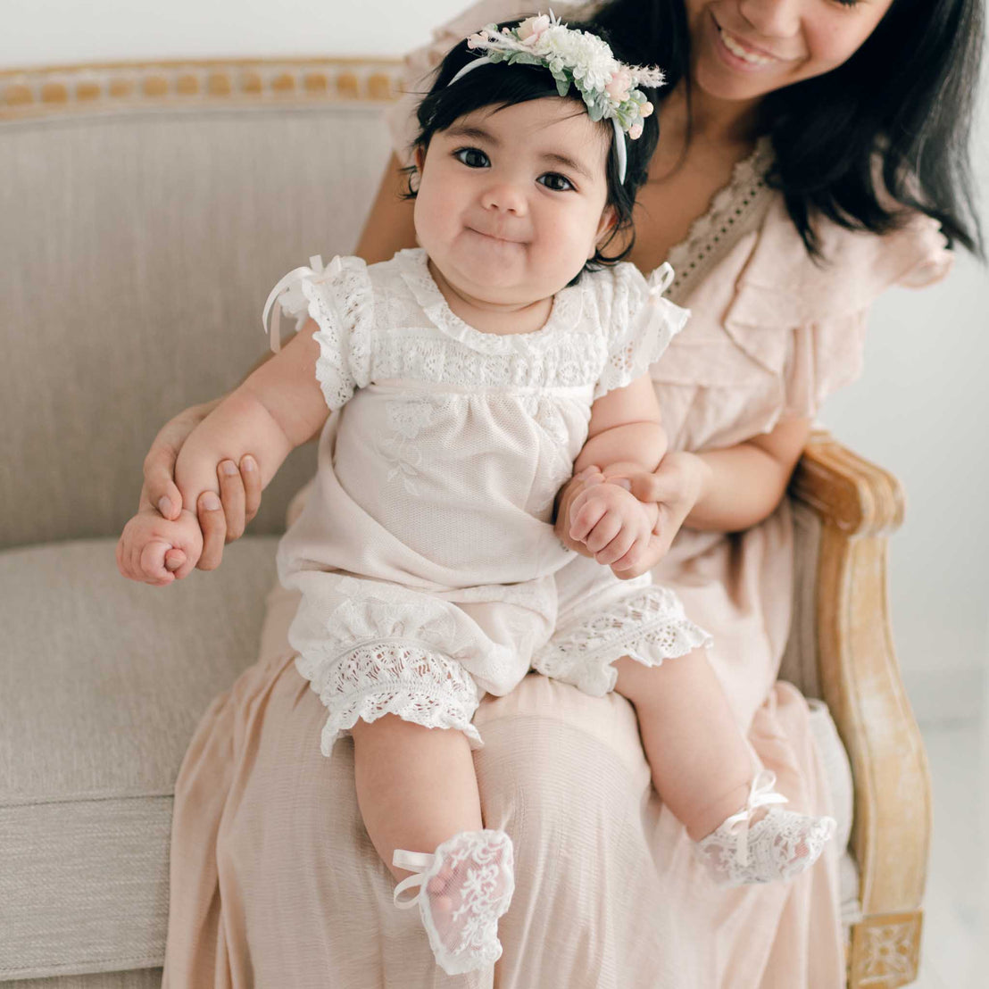 A baby in an ivory lace-trimmed outfit and floral headband sits on a woman's lap, dressed in the Charlotte Convertible Skirt & Romper Set. They are seated on a beige upholstered bench.