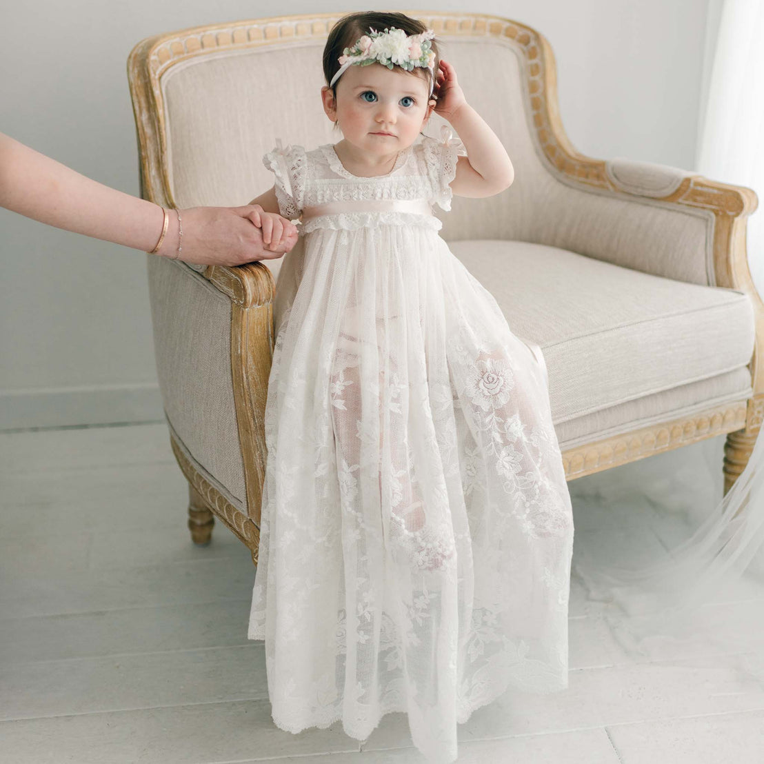 A young child in a white Charlotte Convertible Skirt and floral headband stands on a light-colored armchair, ready for her heirloom christening. An adult's hand gently supports her in the softly hued room, creating a scene of delicate charm and care.