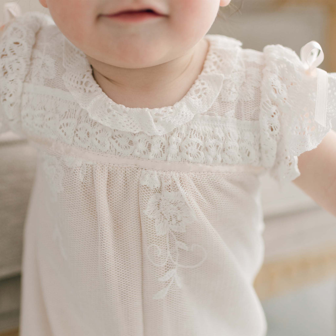 A young child is partially visible, wearing an elegant Charlotte Convertible Skirt & Romper Set that resembles a classic christening gown. The focal point is on the intricate patterns of the skirt, while the child's face softly fades out of frame against a beautifully blurred background.