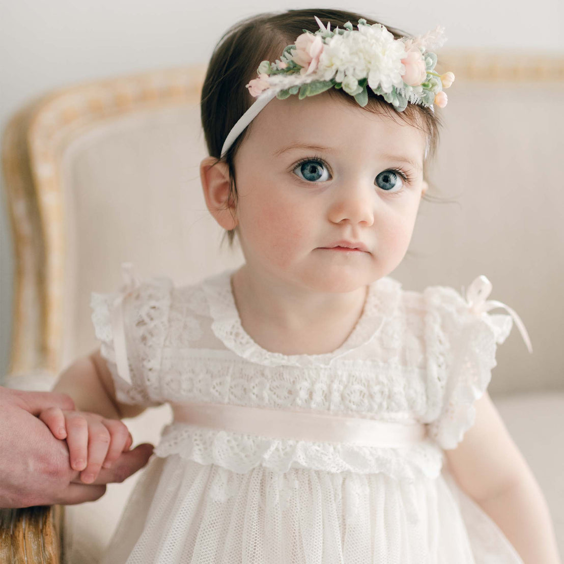 Baby girl wearing the Charlotte Flower Headband, featuring pink and white flowers. She sits on a linen chair holding onto a hand to the side of her.