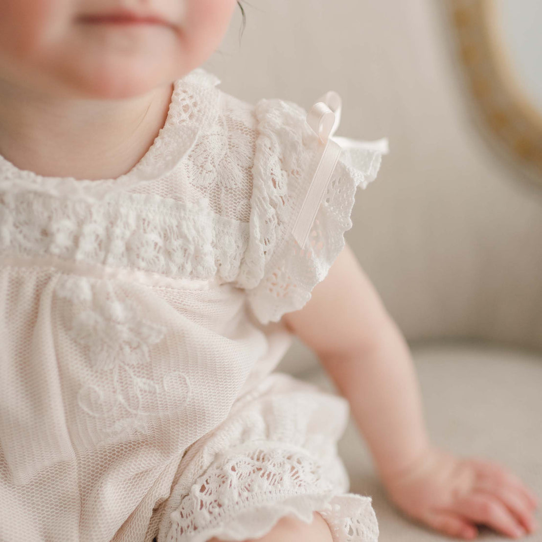 Close-up of a baby in the Charlotte Convertible Skirt & Romper Set, featuring intricate ivory lace with a small ribbon on the shoulder. The baby is partially visible, seated on a soft surface, showcasing the delicate lace details of this exquisite outfit.