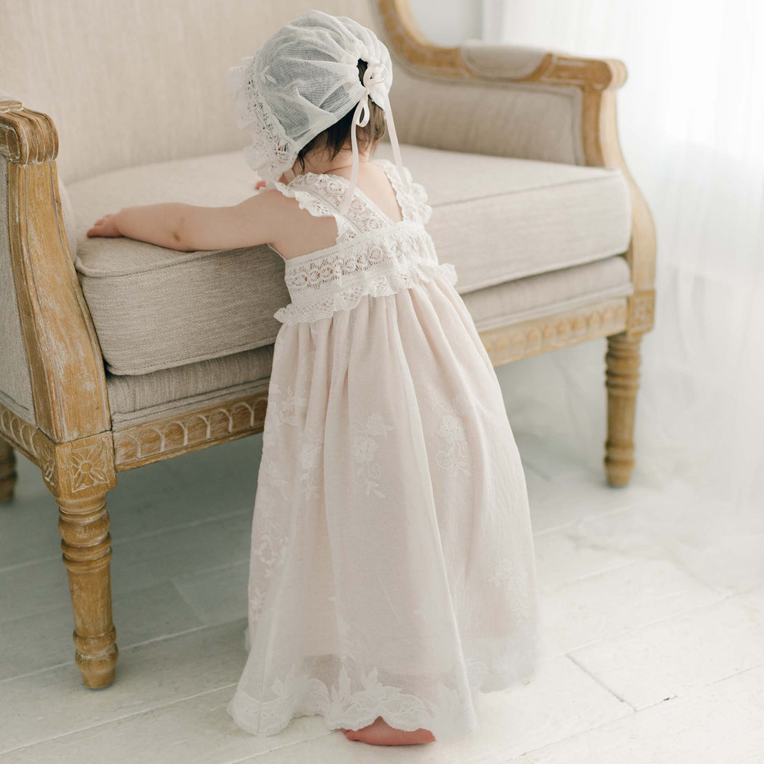 A young child in a Charlotte Christening Gown & Bonnet with rose and vine embroidery stands with her back to the camera, arms outstretched, in a bright, white room.