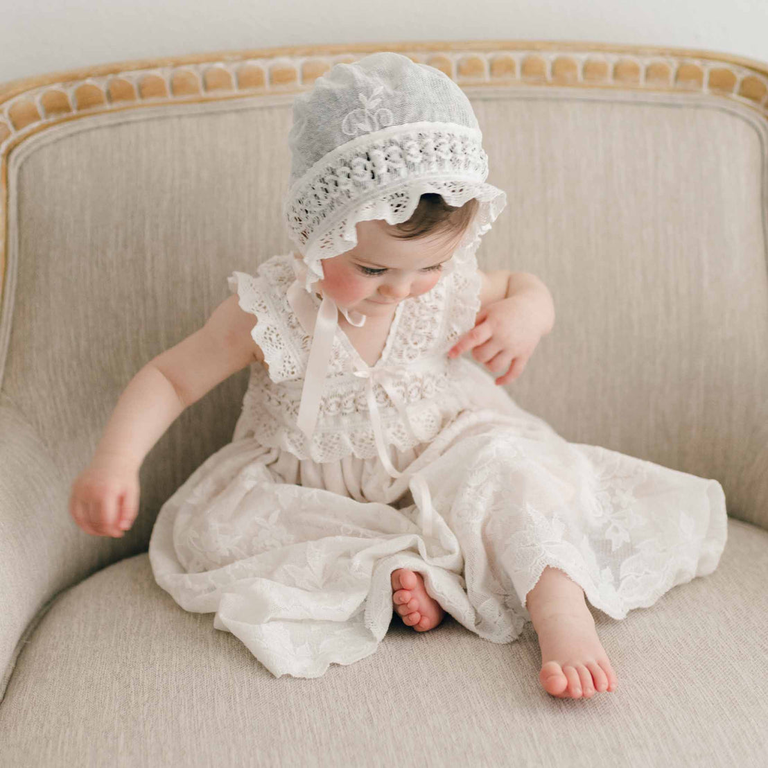 A toddler in a Charlotte Christening Gown & Bonnet sits on an elegant cream-colored chair, looking down thoughtfully. The background is a soft, beige and wood chair.