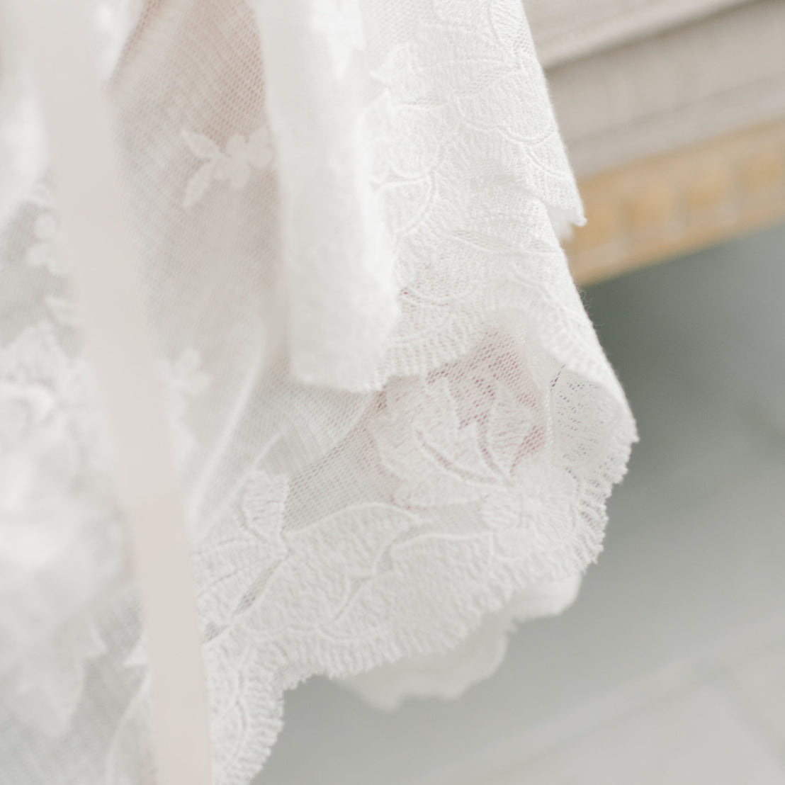 Close-up of delicate Charlotte Christening Gown & Bonnet lace fabric with intricate rose and vine embroidery draped gently over a surface, illuminated by soft natural light.