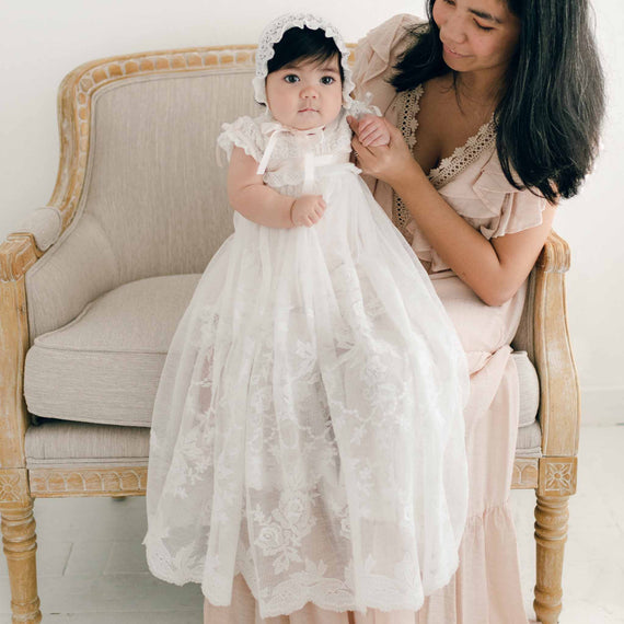 A woman seated on a beige armchair holds a baby dressed in the exquisite Charlotte Convertible Skirt & Romper Set, featuring an ivory lace gown and bonnet. The baby gazes forward as the woman smiles slightly, admiring the child. The indoor setting with its neutral-colored decor perfectly showcases the elegant christening ensemble.