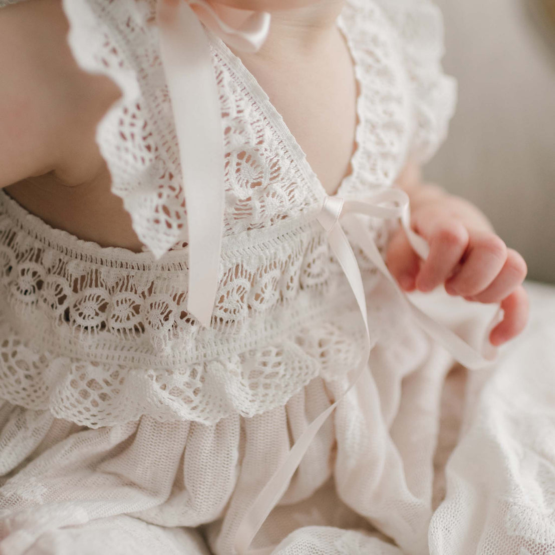 Close-up of a delicate Charlotte Christening Gown & Bonnet with intricate cotton lace details and a soft pink ribbon, displayed on a hanger. The texture of the fabric showcases a fine, detailed pattern.