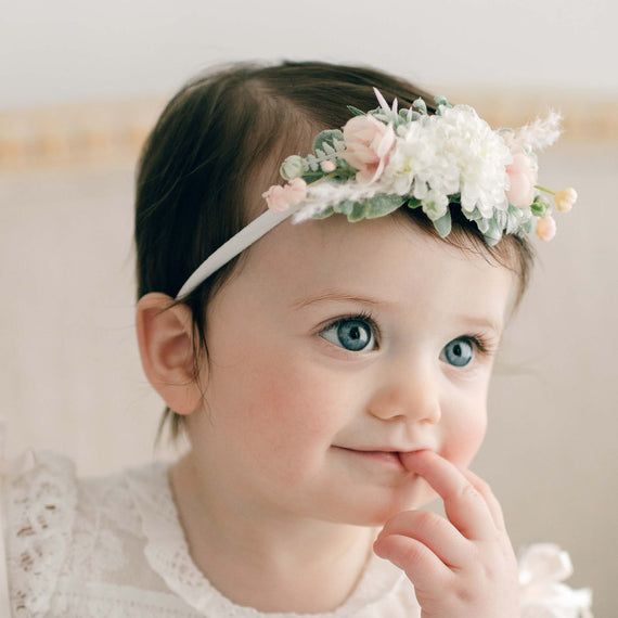 Close-up of a baby girl wearing the Charlotte Flower Headband adorned with pink and white flowers. Her blue eyes are focused off to the side as she gently touches her lips.