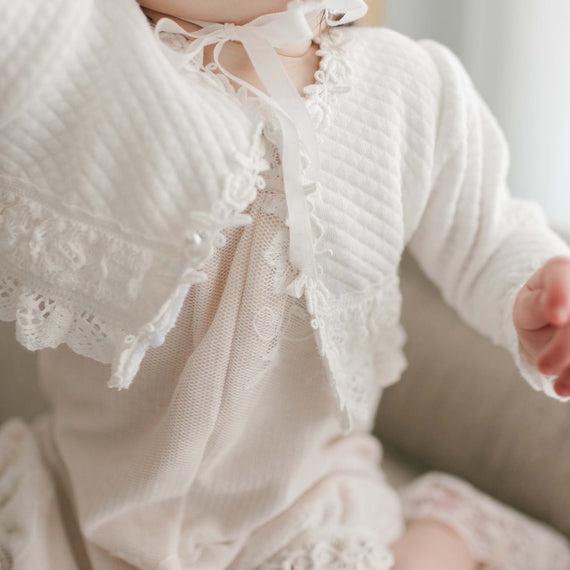 In the image, a baby models the Charlotte Quilted Cotton Sweater, an exquisite white jacket adorned with intricate rose and vine lace details and a ribbon tie. This outfit, made from ivory quilted cotton, boasts delicate embroidery and charming pearl-style buttons. The baby's face is obscured by the softly blurred background.