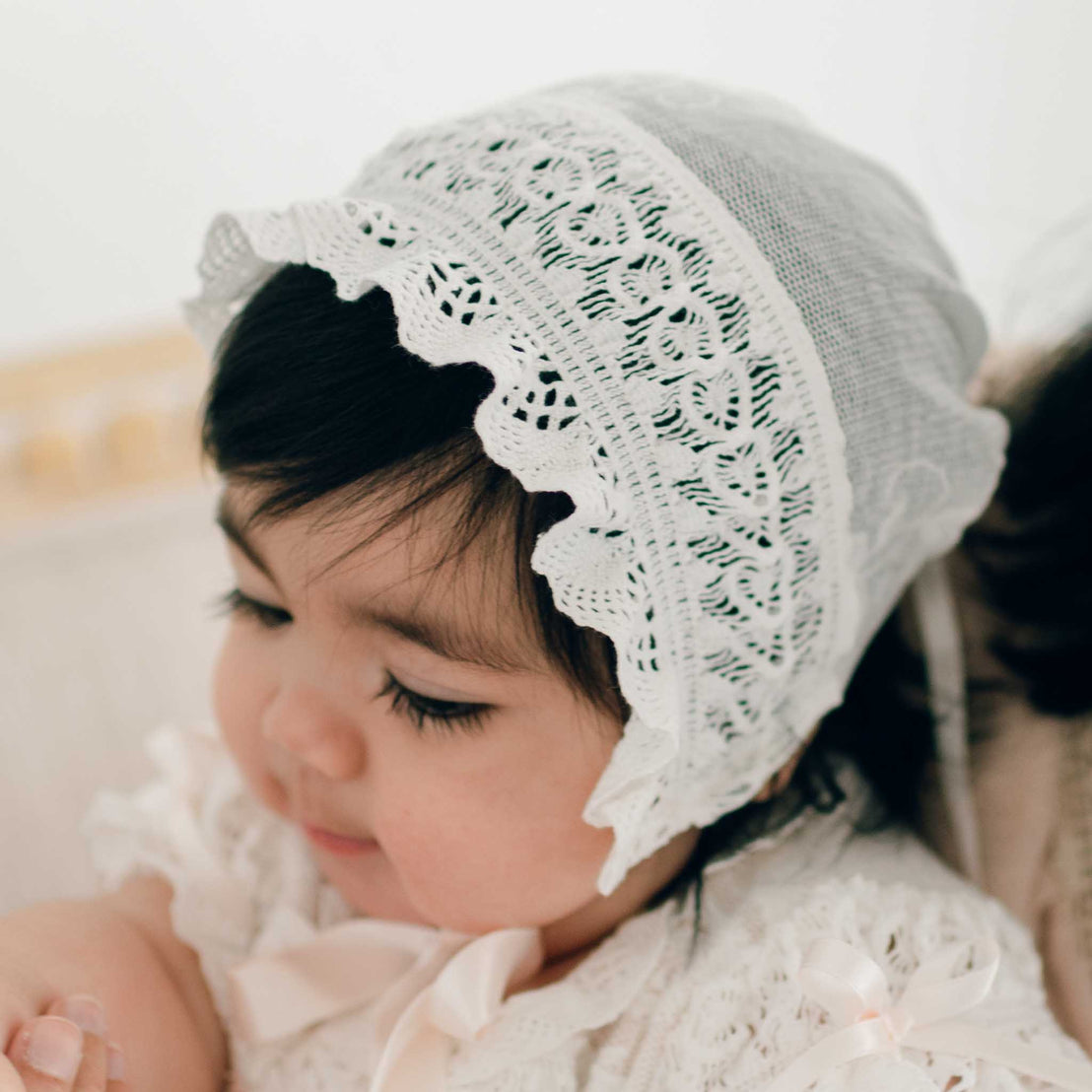 Dressed in the Charlotte Christening Gown & Bonnet, a dark-haired baby graces the indoors under soft lighting. The elegant outfit is adorned with embroidered lace showcasing floral patterns and scalloped edges, making it an ideal baby shower gift.