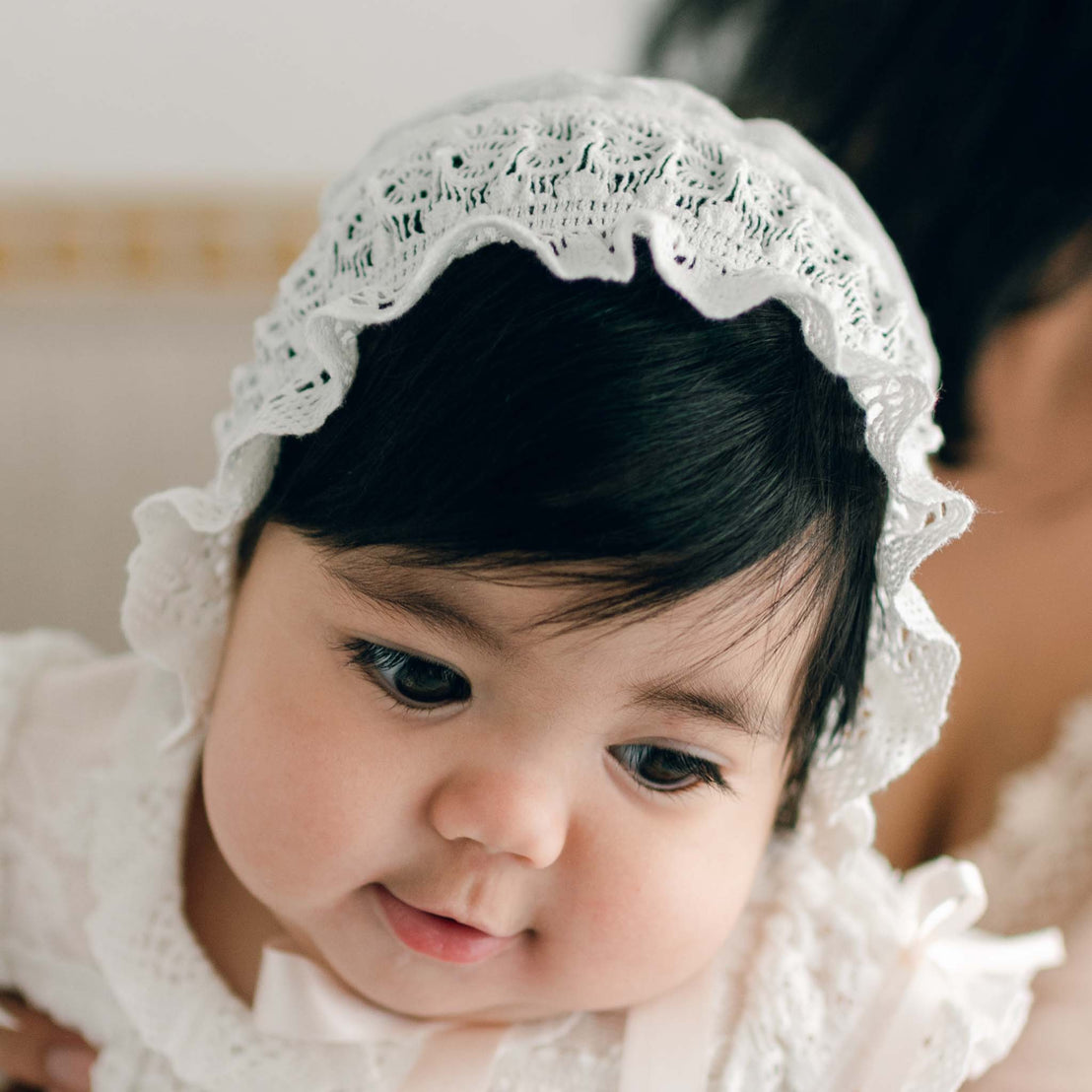 A baby wearing the Charlotte Christening Gown and Bonnet is shown in close-up. The baby has dark hair, a neutral expression, and is looking slightly downwards, with the soft background blurred out of focus.