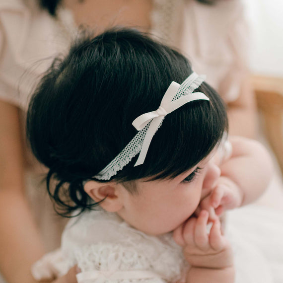 Close-up of a baby girl wearing the Charlotte Pink Silk Ribbon Headband with delicate lace and a soft pink silk bow. Seated on her mother’s lap, she gently holds her hands to her mouth.