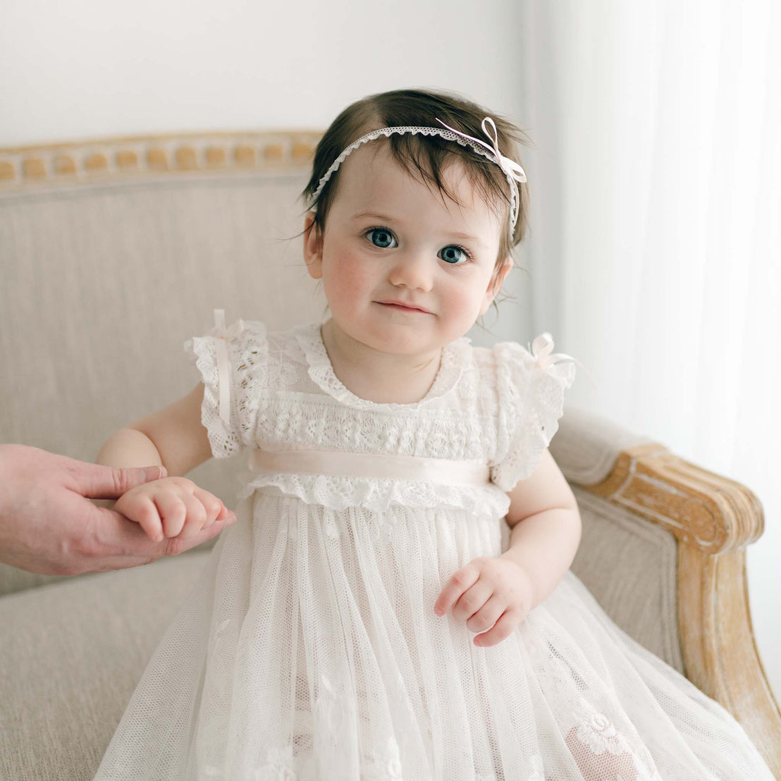 A toddler in the Charlotte Convertible Skirt & Romper Set and headband sits on a light-colored armchair. An adult hand gently holds the child's hand amidst the softly lit natural background.