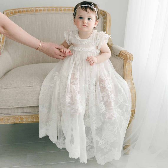 A baby, dressed in the Charlotte Convertible Skirt, sits on an armchair, exuding pure innocence at a baptism ceremony. An adult hand gently holds the baby's hand, with a sheer curtain and light-colored floor in the background, beautifully capturing this heirloom christening moment.