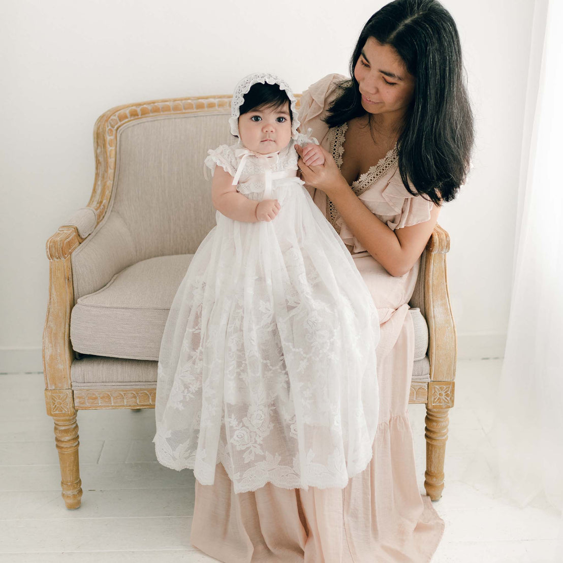 A woman in a beige dress sits on a carved wooden chair, gently holding a baby dressed in the Charlotte Convertible Skirt & Romper Set. The baby stands on her lap, wearing a delicate bonnet against a light-colored, softly lit background.