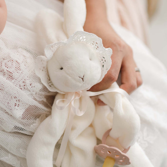 A close-up of the Charlotte Silly Bunny Buddy | Pacifier Holder, a plush white bunny adorned in lace and held by someone. This bunny, ideal for a christening gift, is accessorized with a bonnet and features an attached pacifier clip. The background displays a lace-covered surface.