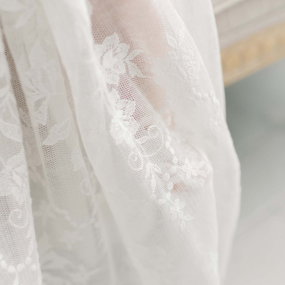 A close-up of the Charlotte Convertible Skirt, showcasing its delicate sheer white fabric adorned with intricate floral lace patterns, reminiscent of an heirloom christening gown. The material partially drapes over a blurred background, evoking an elegant and light atmosphere similar to that of a baptism ceremony.