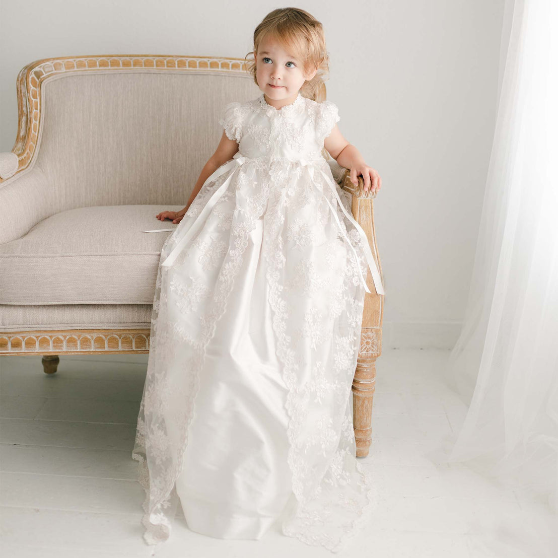 Baby girl without bonnet sitting in chair wearing the full length Christening Gown, the Penelope made of silk and luxurious floral lace.