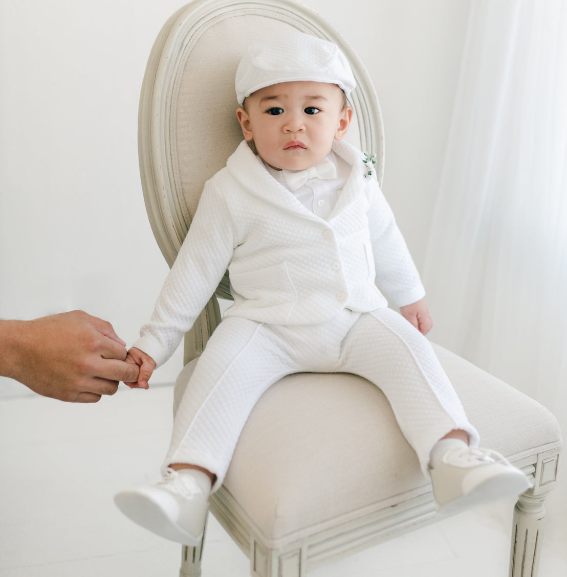 A baby dressed in the Elijah 3-Piece Set, complete with a jacket, pants, and onesie, sits on a beige upholstered chair. An adult hand gently holds the baby’s hand, providing support. The baby is also wearing a matching Elijah Newsboy Cap and White Velvet Bowtie & Boutonniere. The background is a softly lit white room.