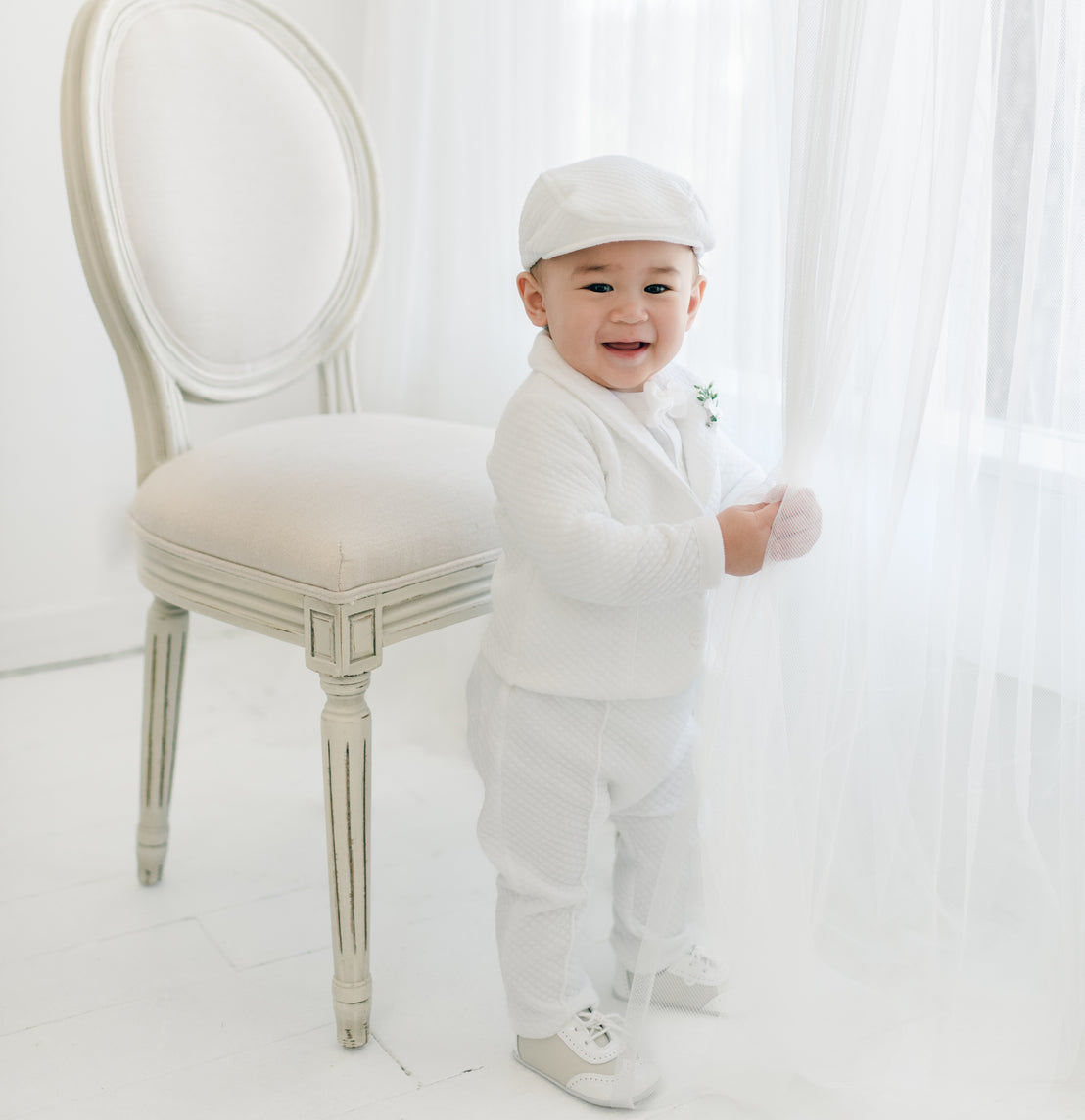 A baby boy stands next to a beige, cushioned chair in a bright room. The baby boy is dressed in the Elijah 3-Piece Set, complete with a jacket, pants, and onesie. Holding onto a sheer white curtain, he smiles at the camera with pure joy. He is also wearing the matching Elijah Newsboy Cap and White Velvet Bowtie & Boutonniere.