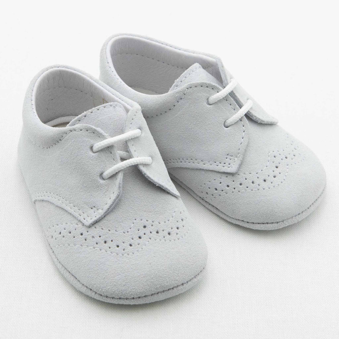 A pair of Dove Grey Suede Shoes from Baby Beau & Belle, featuring white laces and intricate brogue detailing. These handmade baby shoes have a soft sole, making them perfect for infants. They are positioned side by side against a plain white background.